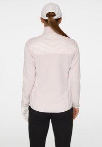 En kvinna står med ryggen mot betraktaren, iförd en ljusrosa jacka med en quiltad axeldesign och svarta byxor, hållande en handske.