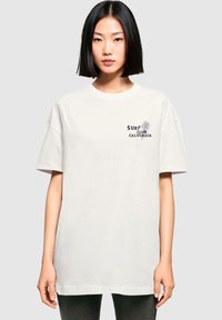 Weißes Baumwoll-T-Shirt mit lockerer Passform, kurzen Ärmeln und einem dunkelbedruckten Grafikaufdruck, der "SURF CLUB CALIFORNIA" mit einem Palmenmotiv zeigt.