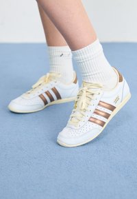 Adidași albi cu dungi bronz, piele texturată, șireturi galbene și detalii perforate. Purtați cu șosete albe cu nervuri pe o suprafață albastră moale.