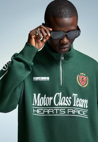 Mand iført en mørkegrøn zip-up sweatshirt med teksten "Motor Class Team Hearts Race", der justerer sorte solbriller mod en ensfarvet baggrund.