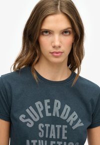 Mørkeblå bomulds T-shirt med korte ærmer, prydet med lys grå tekst, der står "SUPERDRY STATE ATHLETICS" i et buet design.