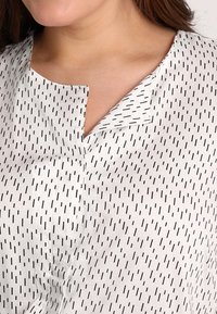 Blouse en tissu blanc avec un motif noir à points, un petit col en V et une coupe décontractée. Le matériau semble lisse avec un léger éclat.