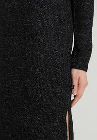 Gebreide jurk in het zwart met een gestructureerd patroon, met zijslits en een label. Toont een close-up van de stof en een hand.