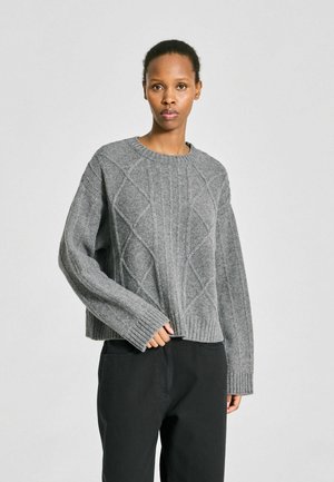 Femme portant un pull torsadé gris et un pantalon noir ample, debout devant un fond clair uni.