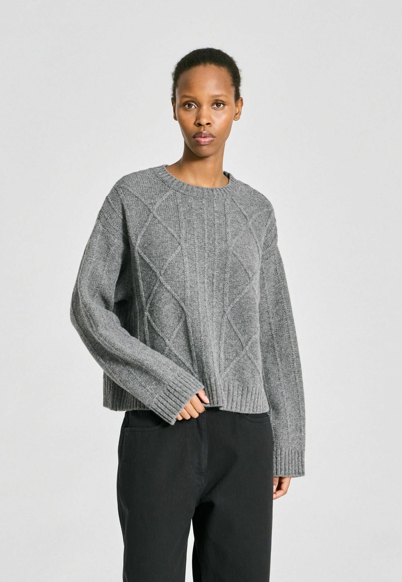 Femme portant un pull torsadé gris et un pantalon noir ample, debout devant un fond clair uni.
