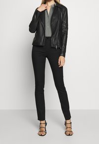 Svart lädermotorcykeljacka med silverdetaljer på dragkedjan, bärs över en grå skjorta med knappar och matchas med svarta åtsittande byxor och sandaletter med remmar.