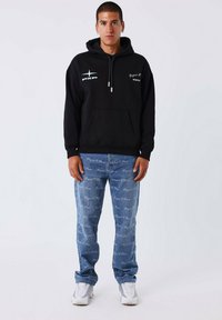 Haut à capuche en coton noir avec cordon de serrage, arborant des impressions de logo. Associé à un jean en denim bleu clair affichant un motif de texte répété. Baskets blanches.