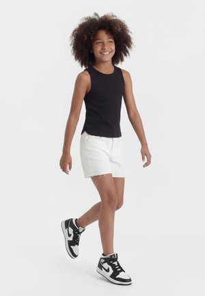 Glimlachend kind met krullend haar dat loopt, gekleed in een mouwloos zwart topje, witte korte broek en zwart-witte hoge sneakers.