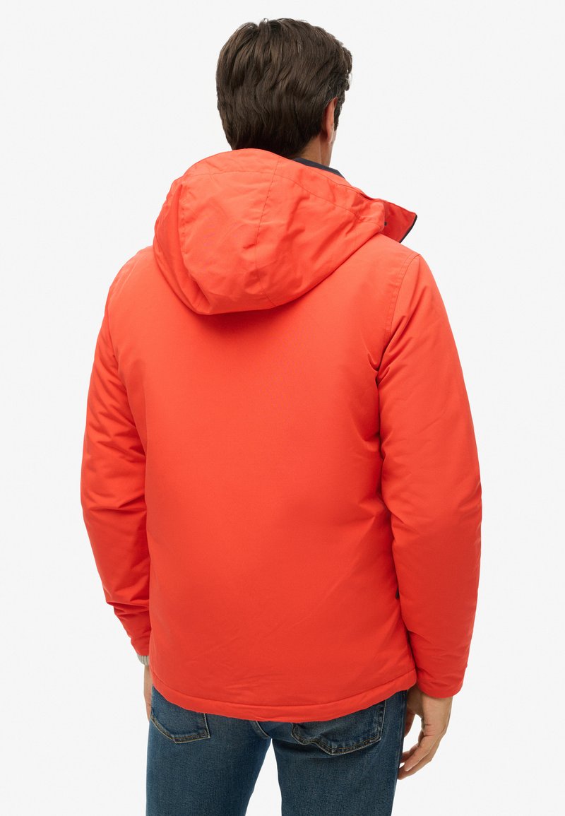 Chaqueta aislante roja con capucha, que presenta una textura suave y puños elásticos. La vista trasera muestra los detalles de las costuras y el diseño ajustado.