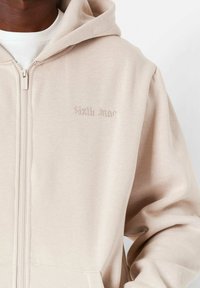 Beige Zip-Hoodie mit weicher Textur, mit einem bestickten Logo auf der Brust und einer Kordelzugkapuze. Mit Kängurutasche.
