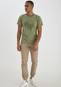 T-shirt en coton vert avec un graphique de palmier, associé à un pantalon jogger beige et des baskets blanches. Coupe décontractée avec des poignets élastiques.