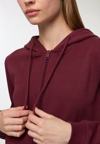 Felpa con zip color marrone bordeaux, dotata di cappuccio con cordino, polsini a coste e una texture morbida. In primo piano sul davanti, mani che regolano la zip.