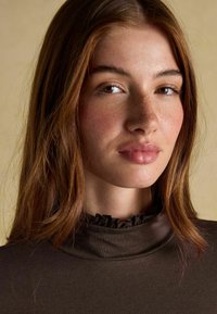 Noor naisel on heledad tedred ja kastanpruunid juuksed, seljas tumepruun kõrge kaelusega särk, millel on volangkaelus, neutraalne taust.