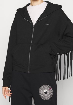 Personne portant un sweat à capuche noir zippé avec des franges aux manches sur une chemise blanche, accompagné d’un pantalon noir avec un graphisme blanc circulaire.
