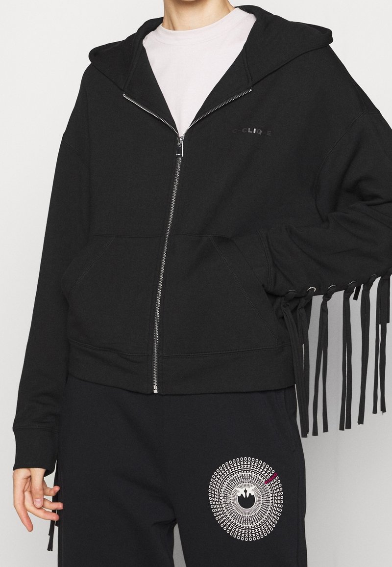 Personne portant un sweat à capuche noir zippé avec des franges aux manches sur une chemise blanche, accompagné d’un pantalon noir avec un graphisme blanc circulaire.