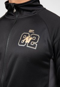 Zwarte zip-up jas met hoge kraag, voorzien van een gouden grafisch ontwerp met het nummer "82" en "EST." aan de voorkant. Glad stoffen textuur.