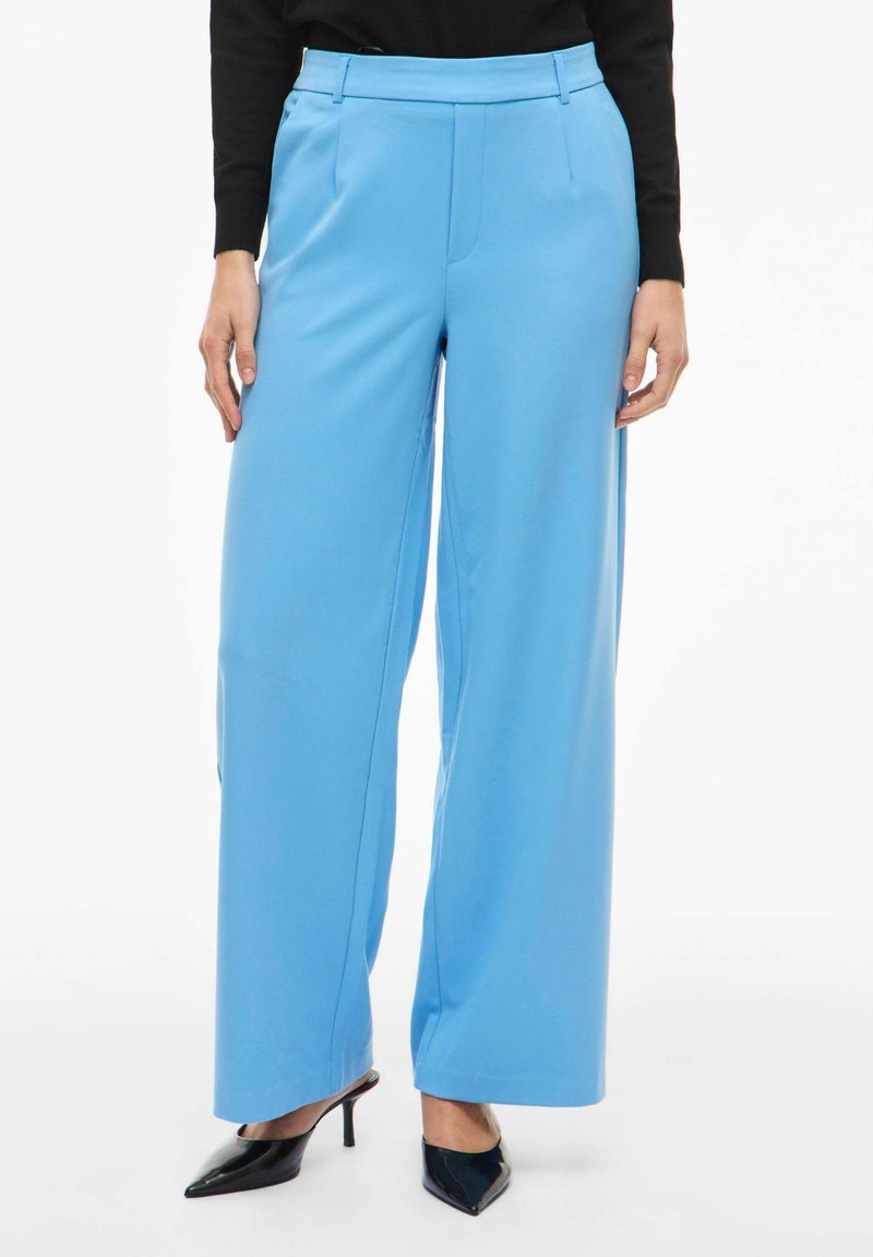 Lyseblå bukser med vide ben og glat tekstur, med fladt frontstykke, bæltestropper og skræddersyet pasform. Kombineret med sorte spidse hæle.