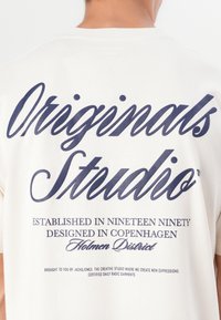 T-shirt en coton blanc avec un texte bleu marine au dos, incluant "Originals Studio", et des détails de design supplémentaires en dessous.