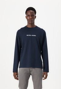 Camisa de manga larga azul marino con "RED BULL RACING" en texto blanco en el pecho, hecha de una tela suave y lisa. Combinada con jeans grises.