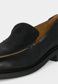 Μαύρο δερμάτινο παπούτσι slip-on με στρογγυλερή μύτη, χαμηλό τακούνι και εσωτερική επένδυση σε ταμπά χρώμα, εμφανίζεται σε λευκό φόντο.