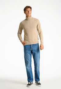 Suéter beige de cuello alto con mangas largas, combinado con jeans de mezclilla azul claro y zapatillas negras. El modelo se encuentra de manera casual contra un fondo simple.