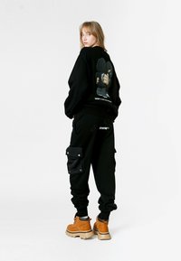 Sudadera negra con estampado gráfico en la parte posterior, combinada con pantalones cargo negros que tienen bolsillos con tapa en la parte frontal. El modelo lleva botas marrones.