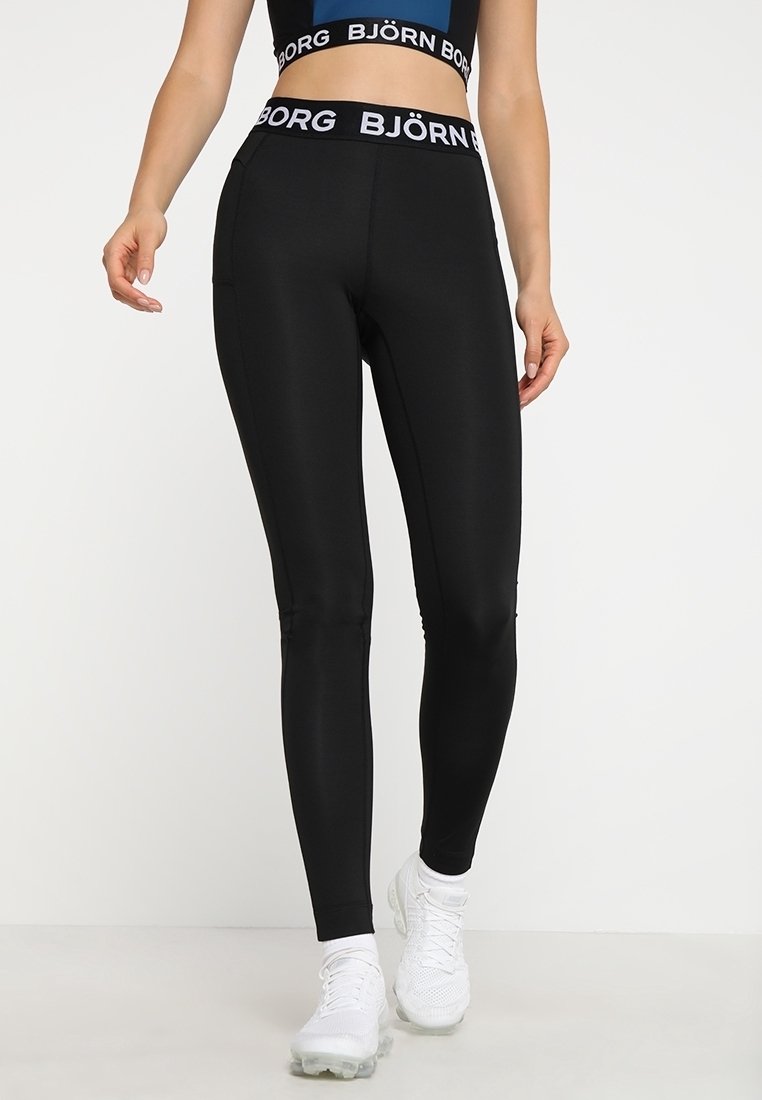 Femme portant des leggings noirs Björn Borg et un soutien-gorge de sport assorti avec des baskets blanches, debout devant un fond clair uni.
