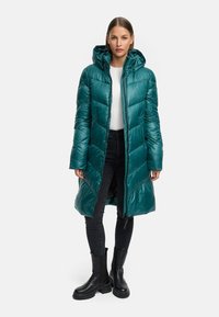 Teal pufferjack met een capuchon, lange snit, gewatteerd ontwerp, ritssluiting aan de voorkant en elastische boorden. Draagt over een witte top en zwarte broek.