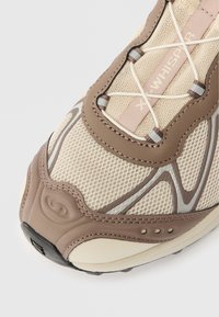 Tenisice s bež mrežastim gornjim dijelom i smešnim akcentima, teksturiranom gumenom potplatom i suprotnim sivim linijama za dodatnu detaljnost.