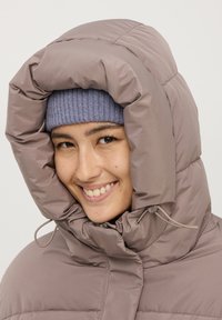 Giacca imbottita con cappuccio in taupe, dotata di un colletto alto e chiusura con coulisse, abbinata a un berretto in maglia a coste di un blu chiaro.