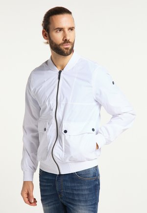 Man with beard wearing white zipped jacket and blue jeans, standing with one hand in pocket, looking to the side.