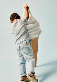 Bambino con una maglietta a righe e jeans sbiaditi che allunga la mano verso un grande cono gelato bianco in schiuma, con una base di cono di cialda marrone texturizzata.