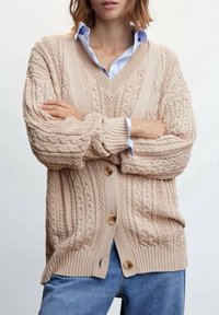 Cardigan en tricot beige avec motifs torsadés, col en V, poignets et ourlet côtelés. Comprend deux boutons et une coupe décontractée. Porté sur une chemise bleu clair.