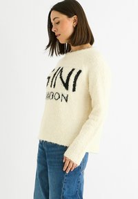 Woman with dark bob hair wears cream fuzzy sweater with black text and blue jeans, standing against a plain white background.