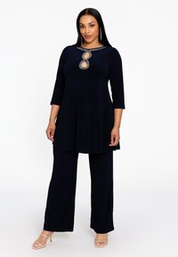 Navyblauwe tuniek met driekwartmouwen, voorzien van een sleutelgat en kraalversiering bij de halslijn. Gecombineerd met bijpassende broek met wijde pijpen.