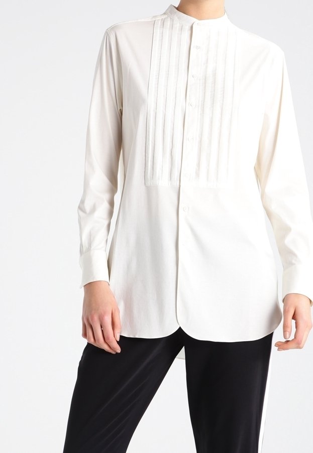 Femme portant une chemise blanche à manches longues avec des plis verticaux sur le devant, associée à un pantalon noir.