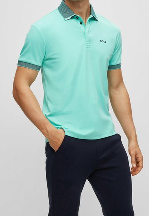 Mann trägt ein mintgrünes Poloshirt mit dunkelgrünem Kragen und Manschetten, marineblaue Hose, Hände in den Taschen, steht vor einfarbigem Hintergrund.