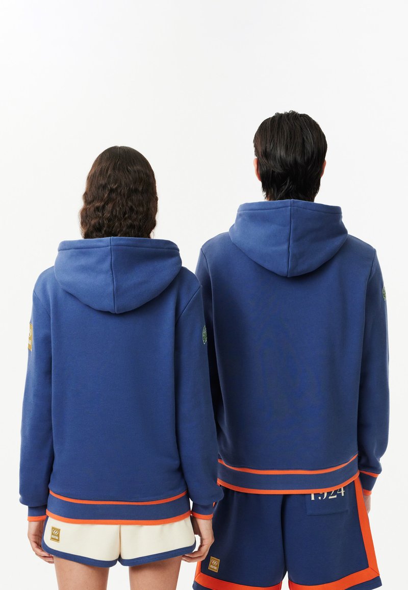 Blaue Kapuzenpullover mit gerippten Bündchen und Säumen, die orangefarbene Akzente aufweisen. Kurze cremefarbene Shorts mit Logopatches, die darunter getragen werden.