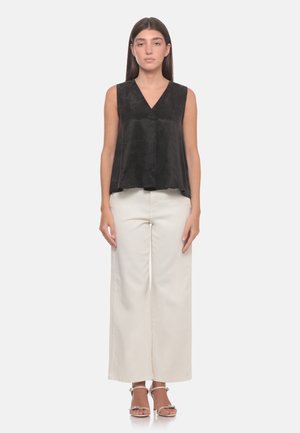 Donna in piedi che indossa un top nero senza maniche, pantaloni bianchi a gamba larga e tacchi aperti su uno sfondo chiaro e uniforme.