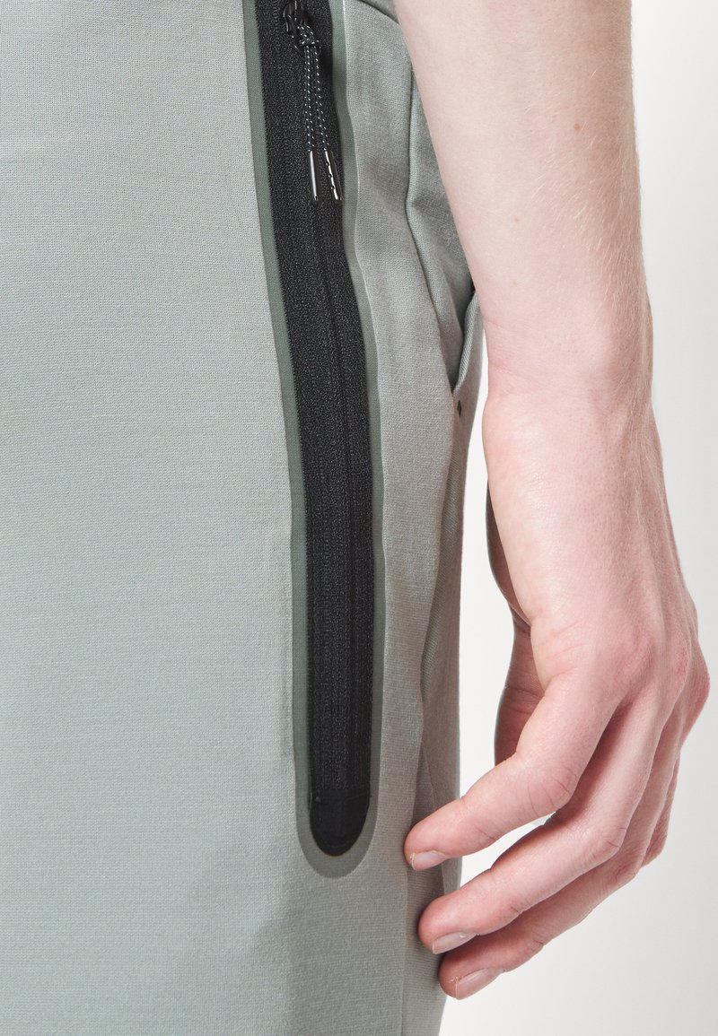 Pantalon de sport gris clair avec une poche zippée noire sur le côté, fabriqué dans un tissu lisse et texturé, au design ajusté.