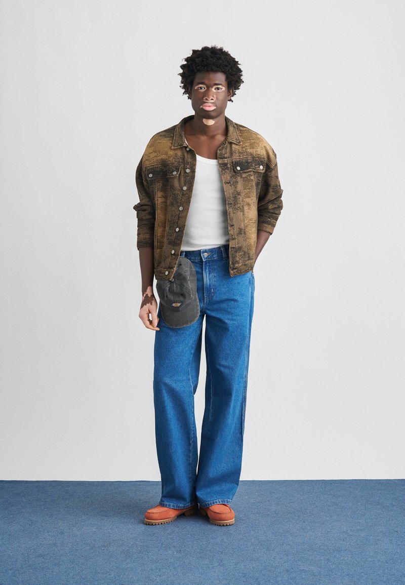 Brown denim jacket over a white tank top, paired with blue wide-leg jeans. Orange-soled shoes and a gray bag accent the outfit.