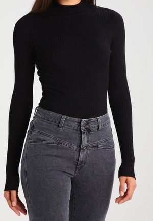 Woman wearing fitted black ribbed long-sleeve top and high-waisted faded black jeans, standing against a plain light background.