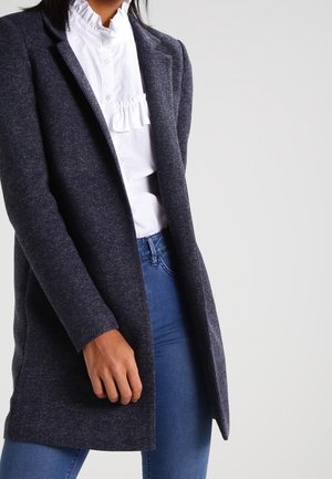 Femme portant un manteau gris foncé ouvert sur un chemisier blanc à volants et un jean bleu, debout devant un fond clair uni.