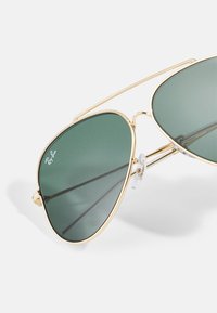 Kultaiset metalliset pilottilasit, joissa on vihreät linssit, kaksoissiltarakenne ja läpinäkyvät nenätyynyt mukavuutta varten. Ray-Ban-logo linssissä.