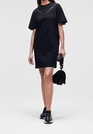 Woman in a black knee-length dress with flutter sleeves, black sneakers, and holding a small black quilted handbag.