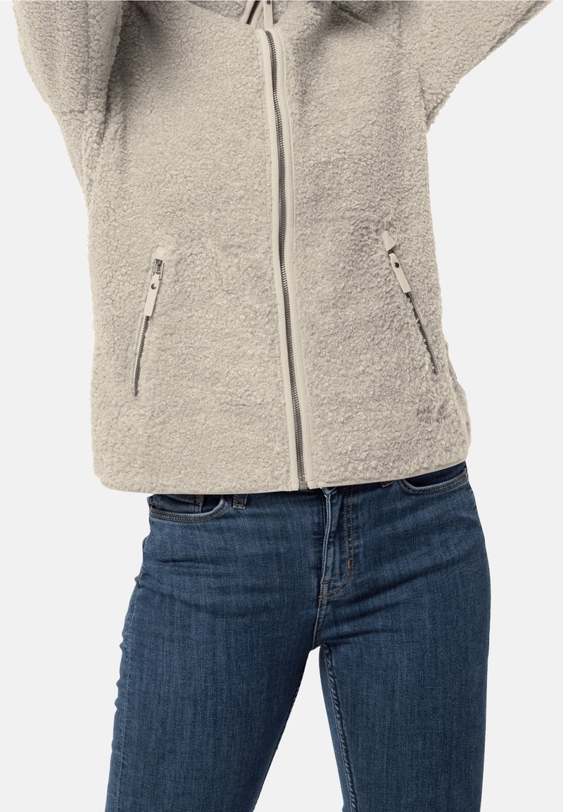 Chaqueta de forro polar color crema con cremallera frontal y dos bolsillos con cremallera, llevada con jeans de mezclilla azules.