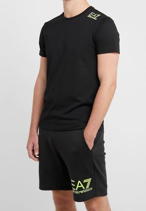 Hombre vistiendo camiseta negra EA7 Emporio Armani y pantalones cortos a juego con logo amarillo neón, de pie con una mano en el bolsillo.