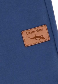 Blauer Stoff mit einer glatten Textur, der ein braunes Lederetikett mit der Prägung "Lagarto Verde" und einer Echsen-Silhouette zeigt.