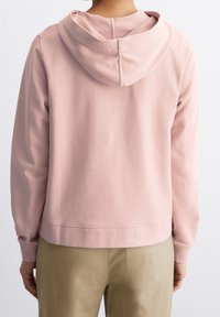 Felpa rosa con una vestibilità comoda, cappuccio con coulisse e polsini a coste. Texture liscia; l'orlo si trova appena sopra la vita. Indossata con pantaloni beige.