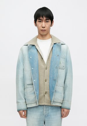 Lyseblå denimjakke med en beige sherpa-krave, lag-på-lag over en beige strikket cardigan med knapper. Bukserne matcher jakken i farve.